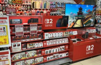 Nintendon suunnitelmat eivät muutu, vaikka PlayStation ja Xbox siirtyvät digitaalisiin peleihin Myymälähylly, jossa on esillä Nintendo Switch 2 -pelikonsoleita, tarvikkeita ja demopiste, jossa pyörii pelikuvaa näytöllä.