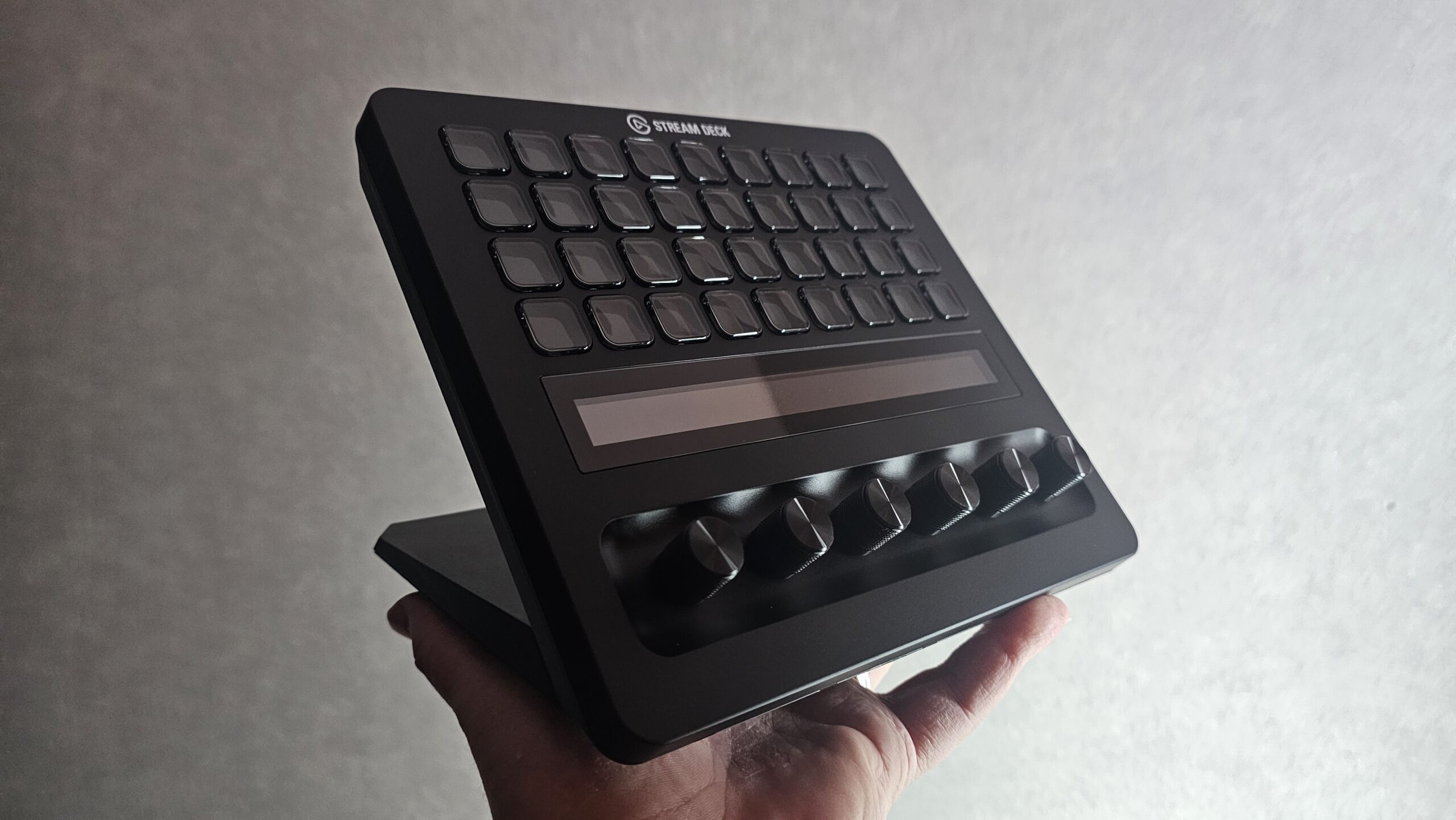 Musta Elgato Stream Deck + XL -ohjainyksikkö kämmenellä pideltynä viistosta kuvakulmasta. Laitteessa on 32 ohjelmoitavaa LCD-näppäintä, pitkänomainen kosketusnäyttöpalkki ja alareunassa kuusi pyöritettävää säätönuppia.