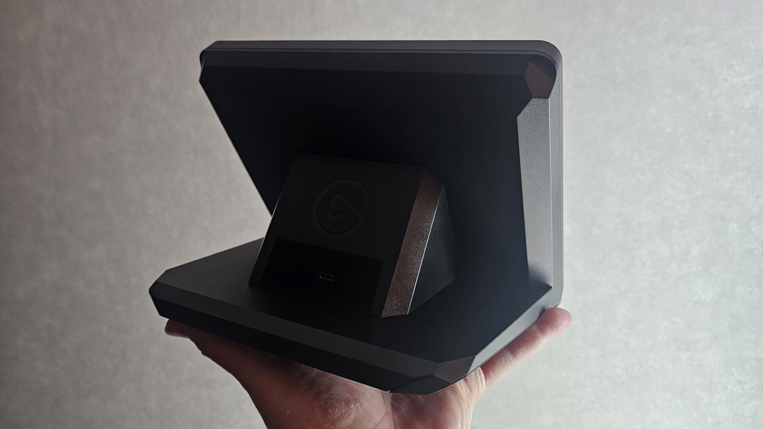 Mustan Elgato Stream Deck + XL -ohjaimen takapuoli kädessä pideltynä. Laitteen jalustassa näkyy kiinteä kulmatuki, valmistajan pyöreä logo sekä syvennykseen sijoitettu USB-C-liitäntäportti.