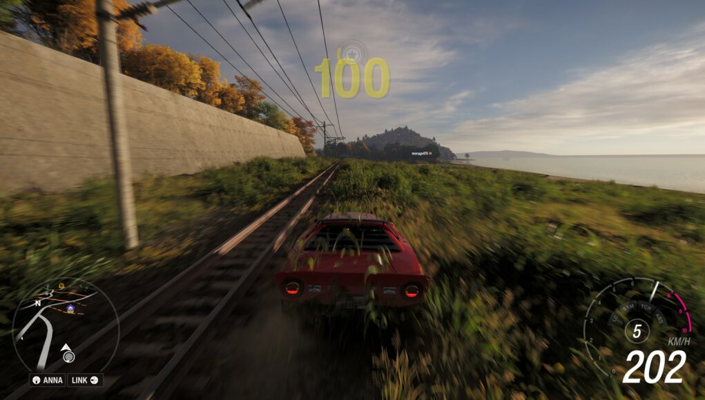 forza-horizon-6-kiskot Pelinäkymä ajopelistä, jossa punainen urheiluauto ajaa vauhdilla rautatiekiskojen vieressä pitkin ruohoista penkereitä rannan ja betoniseinän välissä; näytöllä näkyvät nopeusmittari ja minimappi.
