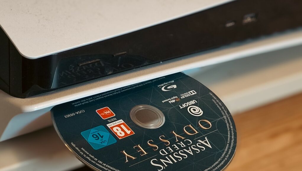 ps5-pelilevy Lähikuva PlayStation-pelikonsolista, jonka levyasemaan on työnnetty Assassin's Creed Odyssey -pelin blu-ray-levy.