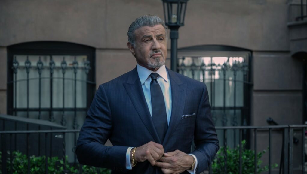 tulsa-king-stallone Muotokuva tummaan pukuun ja solmioon pukeutuneesta näyttelijä Sylvester Stallonesta ulkona rakennuksen edustalla korjailemassa pukuaan.