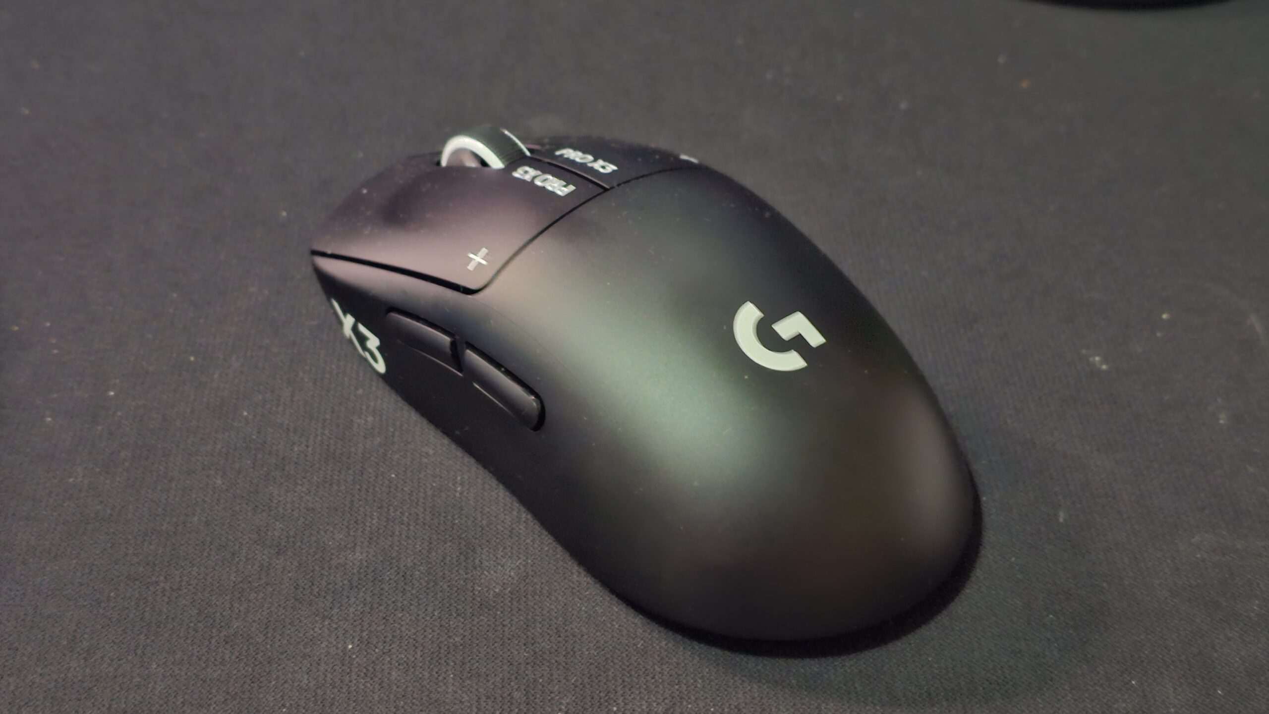 Musta Logitech G Pro X3 Superstrike -pelihiiri kuvattuna viistosti edestä tummalla hiirimatolla. Päänäppäimissä näkyvät valkoiset PRO X3- ja plus-merkinnät sekä kuvioitu rullapyörä, kyljessä kaksi peukalopainiketta ja kämmenosassa valkoinen Logitech G -logo.