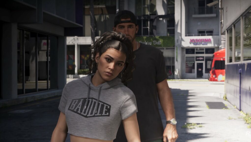 Grand Theft Auto VI -pelin kuvakaappaus, jossa harmaaseen crop top -huppariin pukeutunut Lucia ja lippalakkia pitävä Jason kävelevät ulkona kaupunkimiljöössä päivänvalossa.