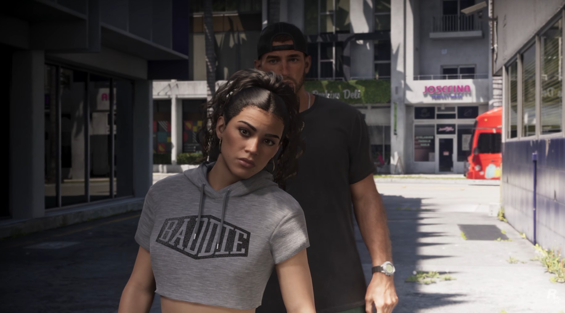 Grand Theft Auto VI -pelin kuvakaappaus, jossa harmaaseen crop top -huppariin pukeutunut Lucia ja lippalakkia pitävä Jason kävelevät ulkona kaupunkimiljöössä päivänvalossa.