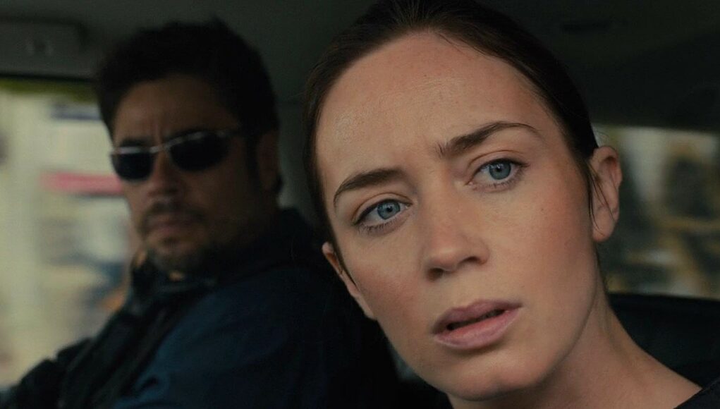 Elokuvakohtaus autosta, jossa etualalla vakavalta näyttävä näyttelijä Emily Blunt katsoo suoraan eteenpäin ja taustalla aurinkolaseihin pukeutunut Benicio del Toro istuu tarkkaavaisena auton matkustamossa.