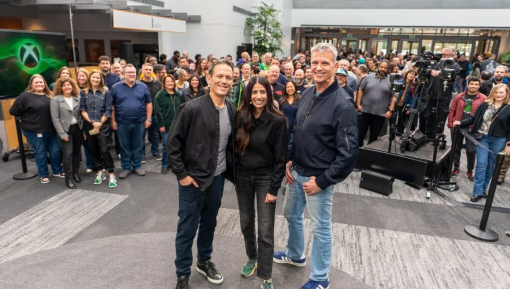 Ryhmäkuva, jossa Xbox-johtajat Matt Booty, Asha Sharma ja Phil Spencer poseeraavat studiossa tai tapahtumatilassa yleisön edessä.